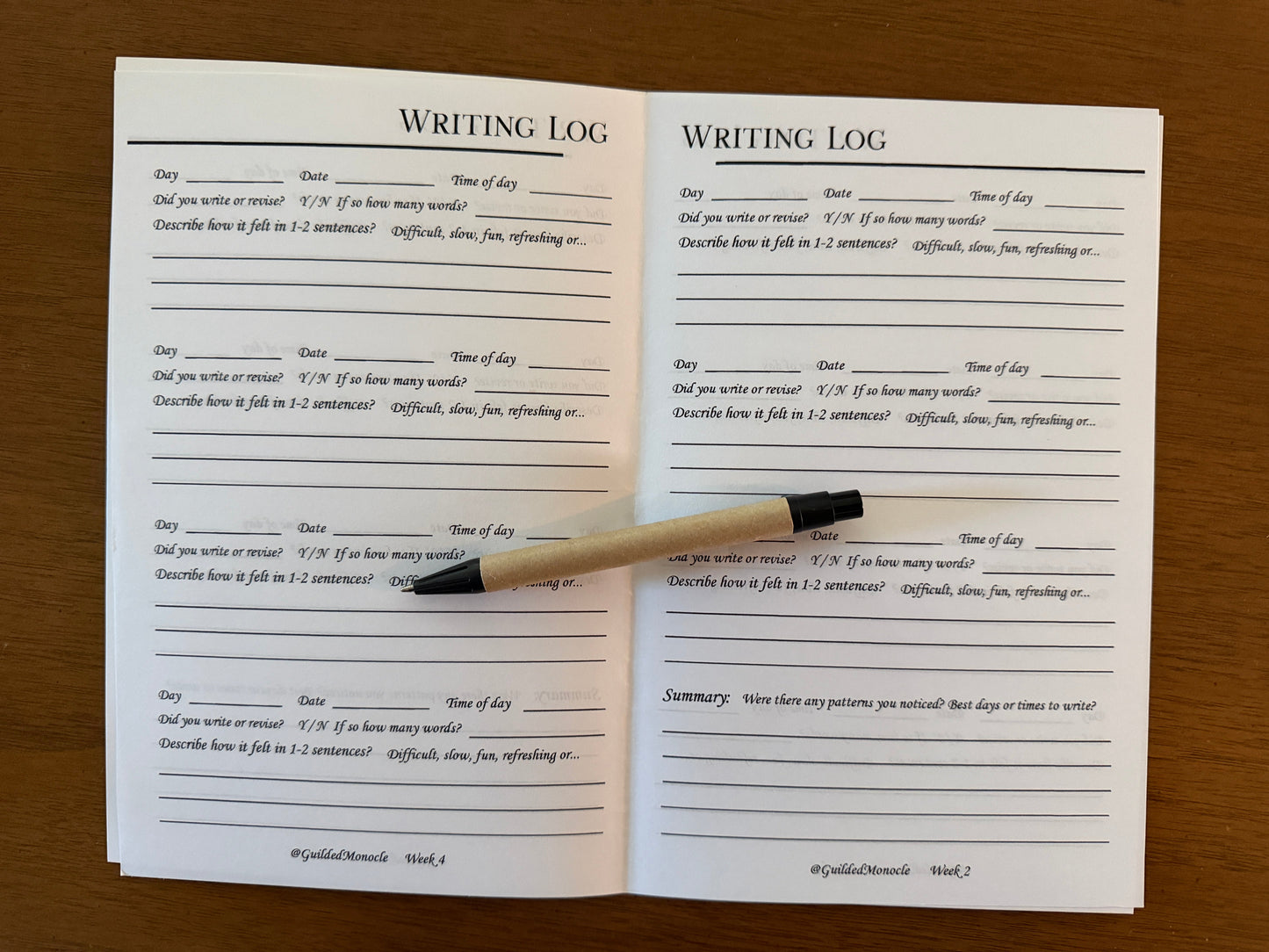 An open writing log booklet with printed prompts and lined writing spaces, placed on a wooden surface with a black and beige pen resting across the pages.