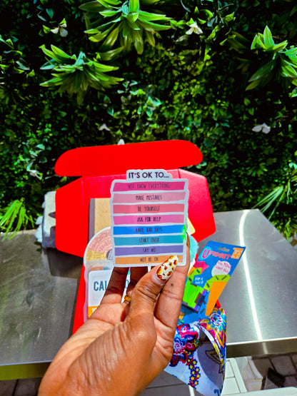 Color swatch sticker of "its okay to" calming held by a hand with a colorful nail design, against a green outdoor background.