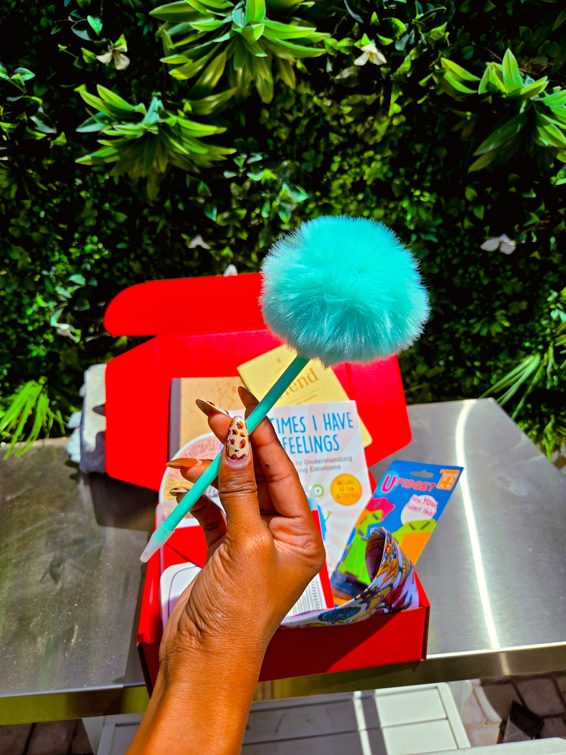 Hand holding a colorful toy with a blue pom-pom, book, and other items on a table outdoors.