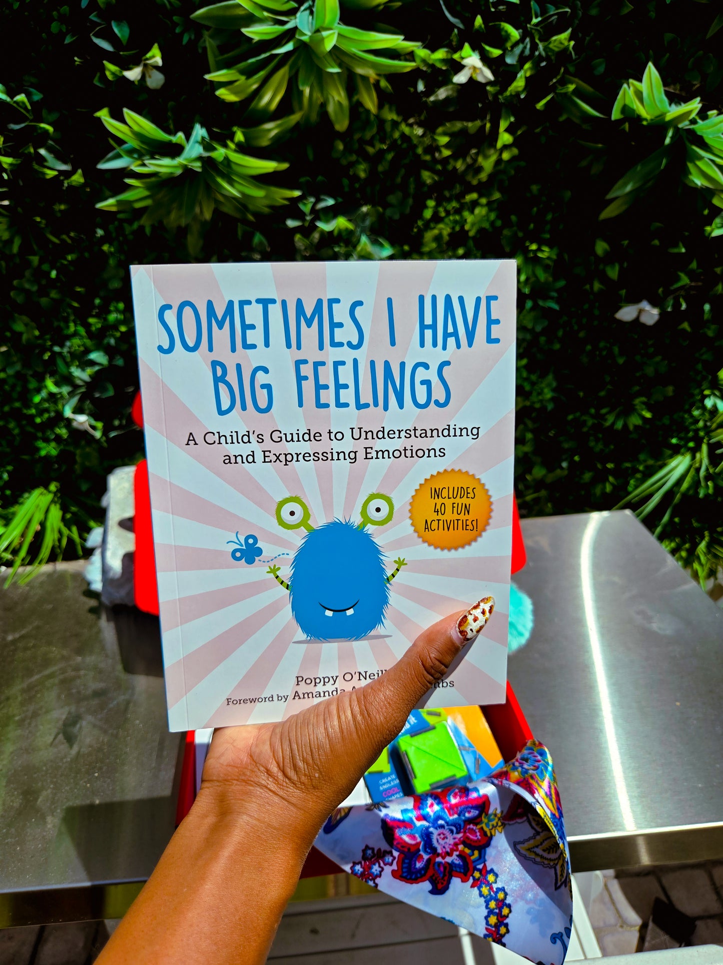 Book titled 'Sometimes I Have Big Feelings' held by a hand with colorful nail polish, against a green outdoor background.