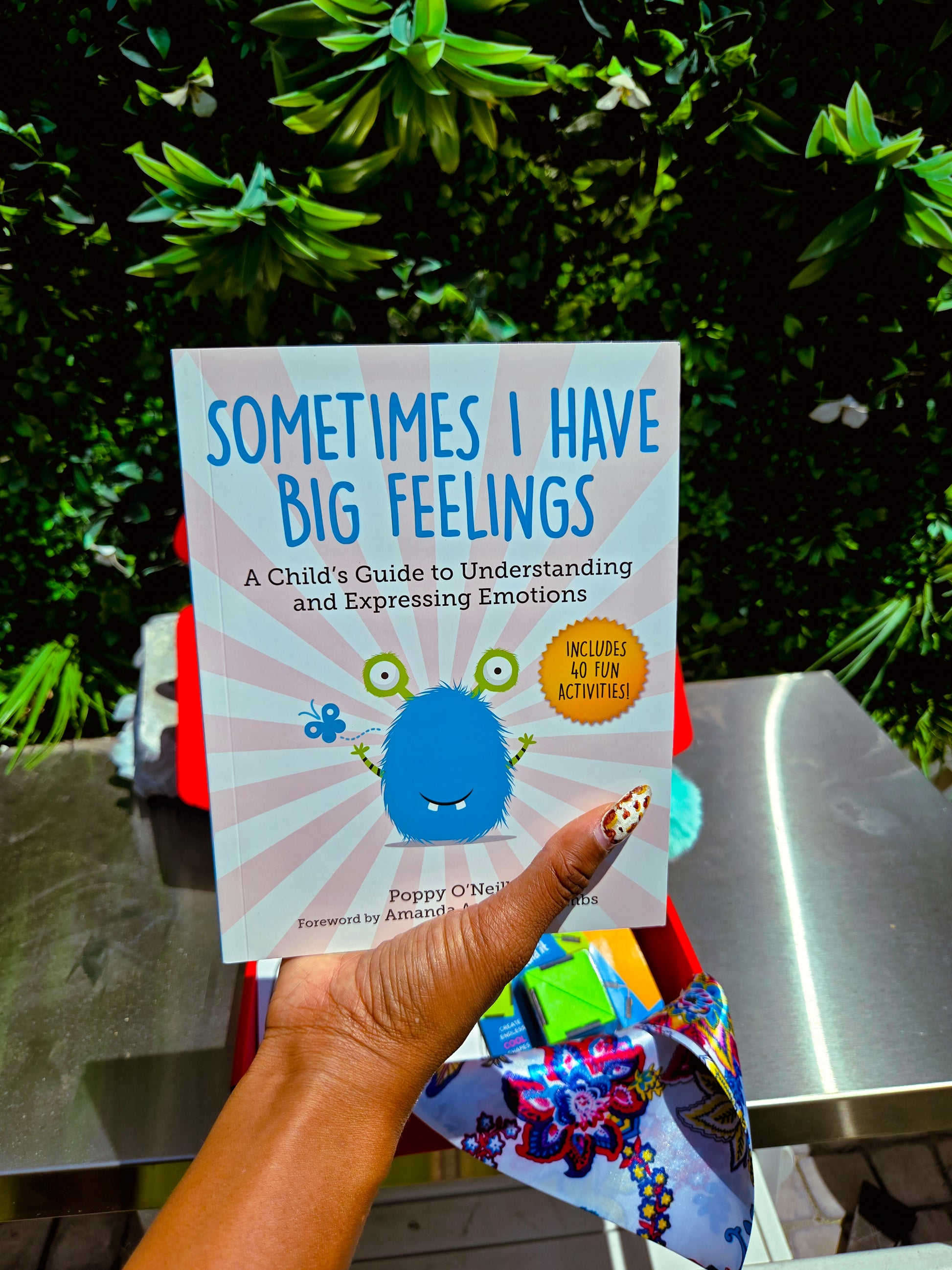 Book titled 'Sometimes I Have Big Feelings' held by a hand with colorful nail polish, against a green outdoor background.